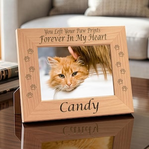 May include: A light brown wooden picture frame with engraved paw prints along the sides. The top text reads You Left Your Paw Prints Forever In My Heart and the bottom text reads Candy. The frame holds a photo of an orange cat.