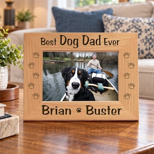May include: A rectangular light wood picture frame featuring the engraved text "Best Dog Dad Ever" at the top and "Brian" with a paw print and "Buster" at the bottom. The frame is decorated with small engraved paw prints along the sides.