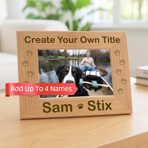 May include: A light-colored wooden picture frame with a horizontal orientation. The frame features engraved paw prints along the sides, the text "Create Your Own Title" at the top, and "Sam Stix" with a paw print at the bottom. A red banner reads "Add Up To 4 Names".