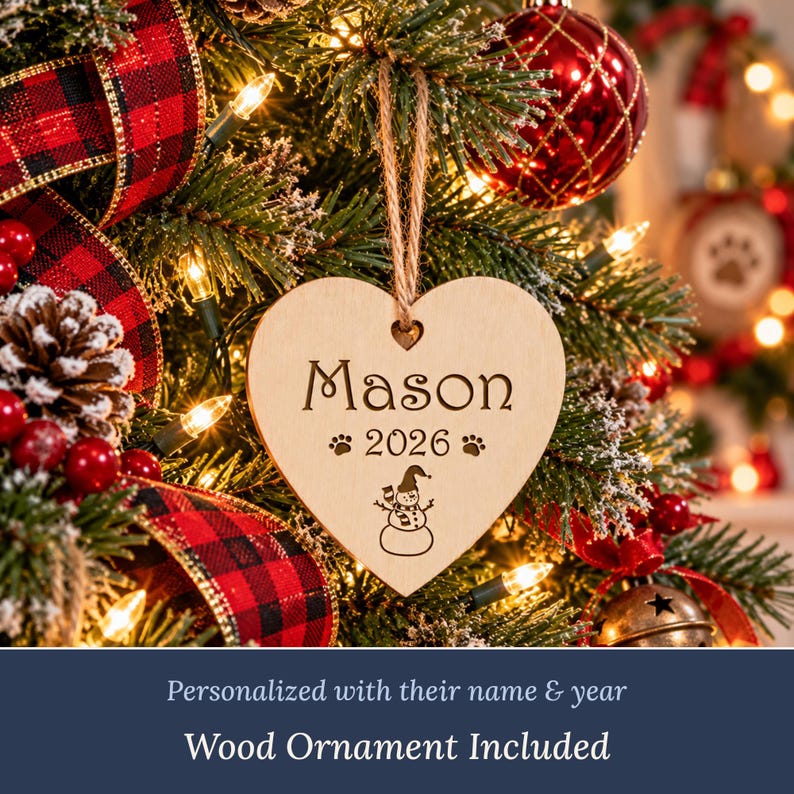 May include: A heart-shaped light wood ornament hanging by a jute twine loop. The ornament is engraved with the name Mason, the year 2026, two paw prints, and a small snowman graphic. The surface is smooth and light-toned wood.