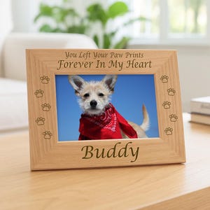 May include: A light-colored wooden picture frame featuring the engraved text "You Left Your Paw Prints Forever In My Heart" at the top and "BUDDY" at the bottom, with small paw print icons along the sides, holding a photo of a dog in a red bandana.