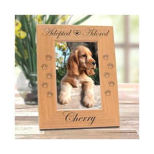 May include: A wooden picture frame with the words "Adopted Adored" and "Cherry" engraved on it. The frame holds a photo of a golden Cocker Spaniel puppy. Paw prints are etched around the frame's border.