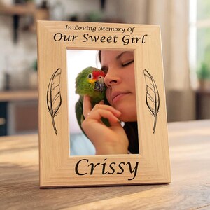 Könnte beinhalten: Ein hellfarbiger Holzbilderrahmen mit dem Text "In Loving Memory Of Our Sweet Girl" und "Crissy". Der Rahmen zeigt ein Bild einer Person mit einem Papagei. Schwarze Federillustrationen befinden sich auf jeder Seite des Rahmens.