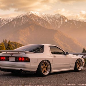 May include: A white sports car with a rear spoiler and gold wheels parked on an asphalt road. The vehicle features red taillights and a sleek body design. The text NijionE is visible in the bottom right corner of the image.