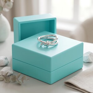May include: A polished silver ring featuring a knotted design resting on top of a square, matte turquoise jewelry box. The ring has a smooth, reflective metallic finish and is centered on the lid of the box, which is open with the lid tilted back.