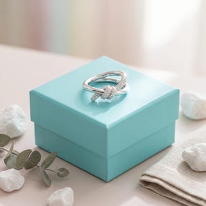 May include: A silver-toned ring featuring a knot design with small clear stones embedded along the surface. The ring is resting on top of a square, bright turquoise gift box with a smooth, matte finish and a lid that sits flush with the base.