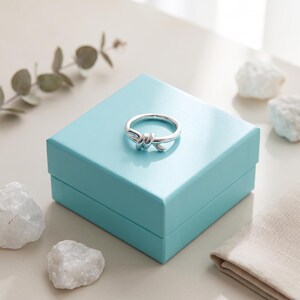 May include: A polished silver ring featuring a central knot design resting on top of a square, light blue gift box. The ring has a smooth, reflective metallic finish and a circular band, presented as a single piece of jewelry on the box lid.