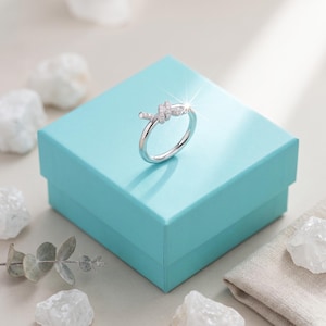 May include: A silver-toned ring featuring a knot-shaped centerpiece adorned with small, clear pavé-set stones. The ring is positioned on top of a square, bright turquoise-colored gift box with a smooth, matte finish and sharp edges.