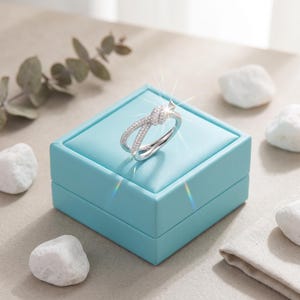 May include: A silver-toned ring featuring a knot design at the center, with the band and knot encrusted with small, clear, sparkling stones. The ring is displayed on top of a square, light blue jewelry box with a smooth, leather-like finish.