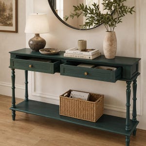May include: A dark green wooden console table with two open drawers, each featuring a small round brass knob. The table has four turned legs and a lower shelf holding a rectangular woven wicker basket. The surface holds a lamp and books.