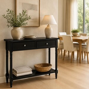 Puede incluir: Una mesa consola rectangular negra con dos cajones y un estante inferior. La mesa tiene patas torneadas y dos pomos plateados. Encima hay un jarrón de cerámica beige con follaje verde, una pila de dos platos y una lámpara beige con pantalla.
