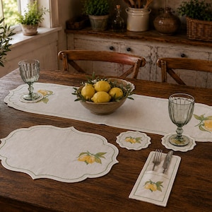 May include: A beige linen table runner and placemat set featuring scalloped edges with green stitching and a printed lemon and leaf motif. The set includes a rectangular cutlery holder, round coasters, and a bowl filled with fresh yellow lemons.