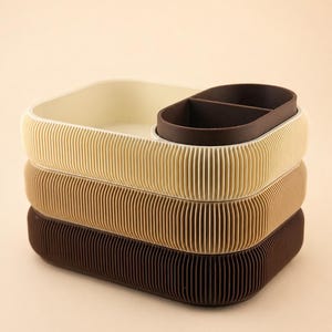May include: A stack of three rectangular desk organizers with rounded corners and vertical ribbed sides. The top organizer is cream with a dark brown divided insert. The middle organizer is tan, and the bottom organizer is dark brown.