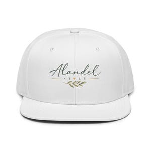 May include: A white snapback cap featuring the text Alandel STYLE in dark green cursive and block lettering. Below the text is a small gold heart and a horizontal line, followed by a green botanical illustration of a leafy branch centered.