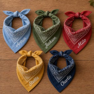 May include: Five triangular pet bandanas in light blue, olive green, red, mustard yellow, and navy blue. Each features a white paisley pattern and a white embroidered name: Arthur, Jackson, Milo, Stella, and Charlie. The bandanas are tied at the top.