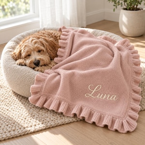 Pode incluir: Uma manta de malha rosa suave com uma borda franzida e o nome Luna bordado em letra cursiva branca. A manta está sobre uma cama redonda e texturizada de cor creme para animais de estimação, onde descansa um cão de pelo encaracolado.