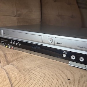 May include: A silver Panasonic DVD and VHS recorder featuring a black front display panel showing 12:00 AM. The device includes a disc tray, a VHS slot, an open/close button, a drive select button, and playback controls with Diga branding.
