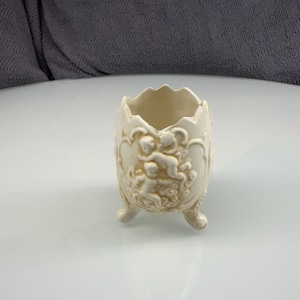 Puede incluir: Un recipiente de cerámica color hueso con forma de huevo y un borde superior dentado que simula una cáscara rota. El exterior presenta un relieve de querubines o putti entre nubes. El contenedor se apoya sobre tres pequeñas patas curvas.