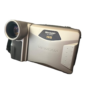 Può includere: Una videocamera Sharp Viewcam VL-AH151 Hi8 color argento con un obiettivo circolare prominente sul lato sinistro, un pannello di controllo nero con il testo Sharp VL-AH151 Hi8 e la parola Viewcam impressa sul corpo del dispositivo.