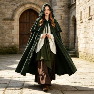 May include: A floor-length dark green hooded cloak with a ruffled trim, worn over a white long-sleeved blouse and a dark green bodice. The outfit includes a long brown skirt with a ruffled hem, creating a layered ensemble with a tie closure.