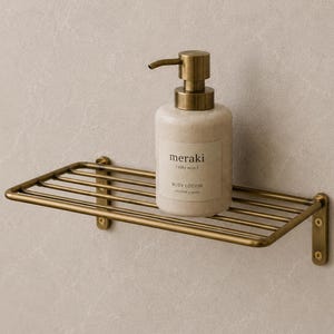Könnte beinhalten: Ein rechteckiges Wandregal aus messingfarbenem Metall mit horizontalen Stangen. Eine beige Pumpflasche mit Meraki Silky Mist Bodylotion steht auf dem Regal. Das Etikett trägt die Aufschrift Meraki, Silky Mist und Body Lotion.