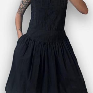 May include: A sleeveless black dress featuring a round neckline, a fitted bodice with a subtle textured pattern, and a wide waistband that transitions into a pleated, knee-length skirt with side pockets for a structured silhouette.
