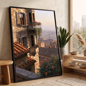 May include: A framed wall art print depicting a sunlit European stone building with terracotta roof tiles, wooden window shutters, and a balcony adorned with red flowers, overlooking a distant town landscape at sunset with warm golden light.