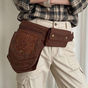 May include: A brown canvas hip bag featuring a large pocket with a floral lace overlay, secured by a flap with metal studs and a central snap button. The bag includes an additional smaller rectangular zippered pouch and a top zipper closure.
