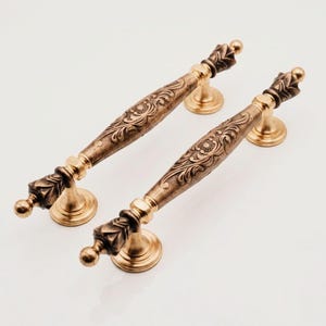 May include: Two elongated cabinet handles featuring a metallic antique brass finish with intricate floral scrollwork patterns. Each handle is supported by two circular mounting bases and terminates in a decorative finial with a leaf-like design.