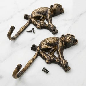 Op de afbeelding: Twee wandhaken van antiek messingkleurig metaal, elk in de vorm van een aap op een tak met een gekrulde staart die de haak vormt. Elk stuk heeft een gedetailleerde vachtstructuur en wordt geleverd met twee kleine zwarte montageschroeven.