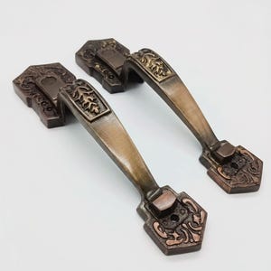 Pode incluir: Dois puxadores de porta a condizer com acabamento em bronze escuro. Cada puxador apresenta uma pega arqueada com um padrão em relevo de folha e placas de montagem hexagonais em ambas as extremidades, decoradas com volutas e orifícios para parafusos.