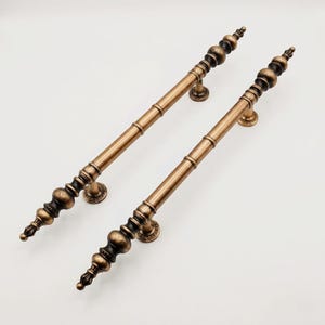 May include: Two long, cylindrical door handles with a metallic antique bronze finish. Each handle features a central rod with decorative, tiered spherical finials at both ends and two circular mounting bases with a textured, engraved edge detail.