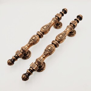 May include: Two antique bronze-colored metal door handles featuring intricate embossed floral patterns, decorative fluted sections, and rounded finials. Each handle is secured by two circular mounting plates with matching ornate detailing.