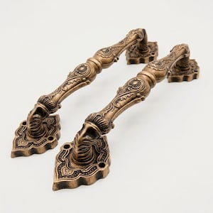 May include: Two antique brass-finished door handles featuring intricate embossed scrollwork and floral patterns. Each handle has a curved grip and is secured by two ornate, diamond-shaped mounting plates with visible screw holes at the corners.