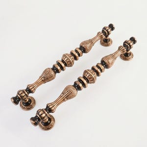 May include: Two long, cylindrical metal door handles featuring a dual-tone finish of antique bronze and dark black. The handles have a repeating pattern of rounded, ribbed, and leaf-like decorative elements, each mounted on two circular bases.
