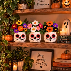 May include: Three crocheted pots shaped like white sugar skulls with black eye sockets and stitched mouths, each filled with a vibrant bouquet of colorful crocheted flowers in shades of yellow, orange, pink, purple, and black with green leaves.