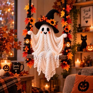Op de afbeelding: Een gehaakte witte spookfiguur met een zwarte heksenhoed versierd met oranje bloemen en bessen, zittend op een schommel versierd met herfstbladeren. Het spook heeft zwarte ogen en een glimlach en houdt twee kleine zwarte lantaarns aan de touwen.