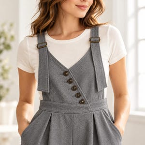 Op de afbeelding: Een grijze mouwloze overgooier met een V-hals, een asymmetrische knoopsluiting, brede schouderbanden met bronzen metalen gespen en een geplooide taille, gedragen over een effen wit T-shirt met korte mouwen en een ronde hals.