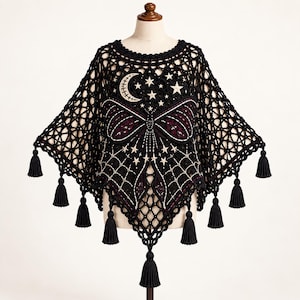May include: A black crochet poncho featuring an intricate openwork pattern with a central embroidered moth design, a crescent moon, and multiple stars. The garment is finished with a round neckline and several black tassels along the hem.
