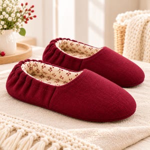 May include: A pair of deep red slippers featuring a gathered elastic collar and a cream-colored interior lining decorated with a small, repeating brown floral pattern. The exterior has a textured fabric finish with a rounded toe design.