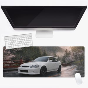 May include: A rectangular desk mat featuring a high-resolution print of a white hatchback car parked on a wet asphalt road. The scene includes a red wooden railing, lush green trees, and blooming cherry blossom trees along a mountain path.