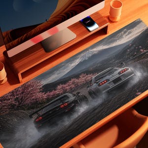 Pode incluir: Um tapete de secretária retangular com um gráfico impresso de dois carros desportivos a fazer drift numa estrada escura. A cena inclui cerejeiras em flor cor-de-rosa e uma montanha ao fundo. Os carros são vistos de trás com luzes traseiras vermelhas.