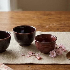 May include: Three dark maroon ceramic bowls with a ribbed texture on the exterior. The bowls have a glossy finish and a rounded shape. Small light pink cherry blossom petals are scattered near the bowls, and a small wooden scoop is placed nearby.