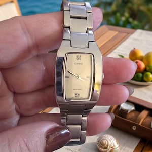 May include: A silver-tone Casio wristwatch with a rectangular case and a matching metal link bracelet. The watch features a champagne-colored dial with silver hands, a Casio logo, and two small decorative accents at the top and bottom positions.
