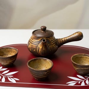 Puede incluir: Una tetera de cerámica marrón con asa lateral y tapa a juego, acompañada de tres tazas de té pequeñas y redondas de cerámica. La tetera y las tazas presentan un esmalte texturizado en tonos marrón y beige con estrías horizontales.