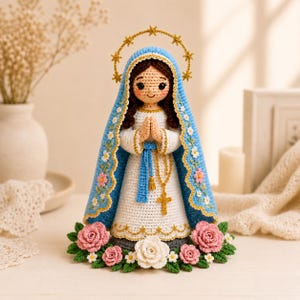 May include: A crocheted figurine of the Virgin Mary wearing a white dress and a blue mantle with floral embroidery. The figure has dark hair, a gold halo with stars, and hands clasped in prayer with a gold rosary. It is surrounded by pink and white roses.