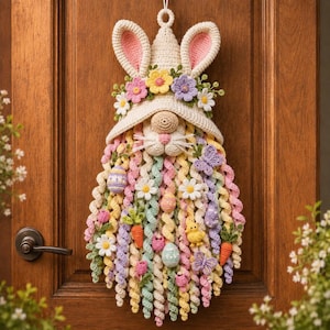 Op de afbeelding: Een gehaakte kabouter-wandhanger met een crèmekleurige muts met roze gevoerde konijnenoren, versierd met pastelkleurige bloemen. De kabouter heeft een lange, krullende baard in roze, geel, groen en blauw, gedecoreerd met kleine gehaakte eitjes, wortels en vlinders.