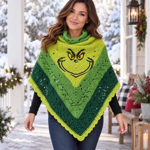 May include: A crochet poncho featuring a gradient of lime green to dark forest green with a scalloped edge. The center displays a black outline of a character face with yellow eyes and a wide grin. The garment has a cowl neck and a V-shaped hemline.