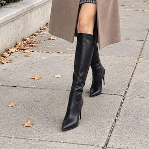 May include: A long tan wool-blend coat with notched lapels and two black buttons, worn over a black turtleneck jumper and a black and white tweed mini skirt. The outfit includes black knee-high stiletto boots and a black shoulder bag.