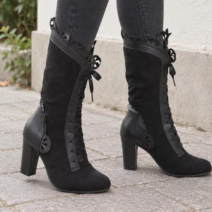 May include: Mid-calf black boots featuring a suede-like texture, a lace-up front with black ribbons, and a block heel. The boots are accented with decorative scalloped trim along the collar and side panels, and include a smooth leather-like heel counter.